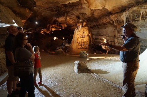 A tour guide explains cave formations to a small group of visitors in a dimly lit cave. Stalactites are visible above.