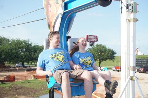 Two people in matching blue shirts enjoy a thrill ride, laughing and smiling as they prepare for the experience.