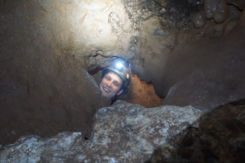 A person with a headlamp smiles while peering out from a narrow cave opening.