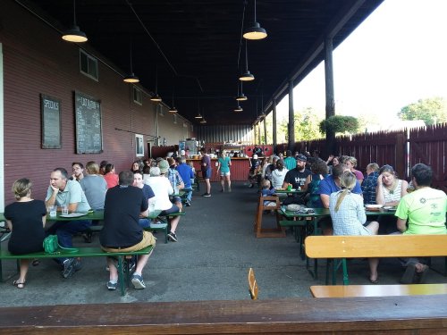 A bustling outdoor dining area with people seated at long tables, enjoying food and drinks under a shaded roof.