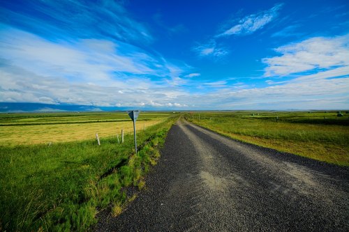 A gravel road stretches through lush green fields under a bright blue sky with scattered clouds.