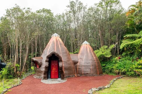 A unique, volcanic-shaped house with a red door, surrounded by lush greenery and trees.