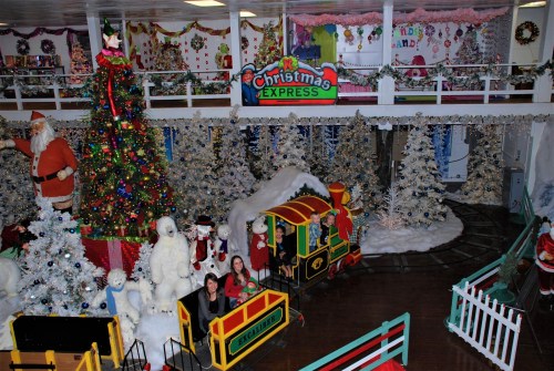 A festive indoor scene with a large Christmas tree, decorations, and a train ride, featuring Santa and snowy displays.