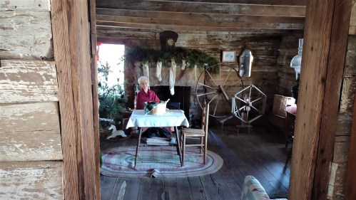 An elderly person sits at a table in a rustic cabin, surrounded by holiday decorations and vintage furnishings.