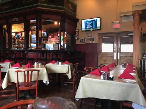Interior of a restaurant with empty tables set with red napkins, a bar area, and a TV displaying sports.