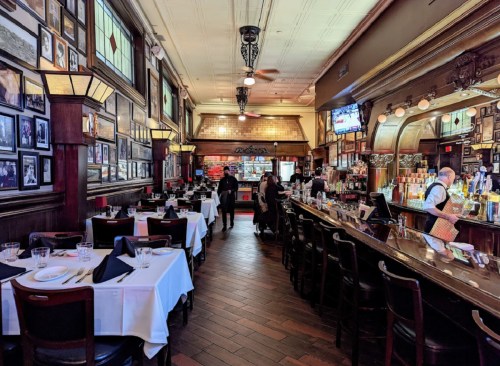 Elegant restaurant interior with wooden decor, tables set for dining, and a bar area in the background.