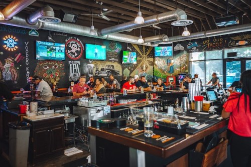 A lively bar scene with patrons at the counter, colorful murals on the walls, and multiple TVs showing sports.