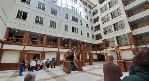 Interior view of a spacious atrium with wooden accents, a staircase, and people gathered on the ground floor.