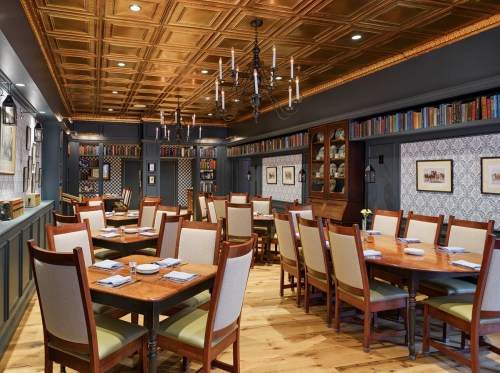 Elegant dining room with wooden tables, chairs, and a coffered ceiling, featuring bookshelves and decorative wall art.
