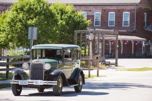 A vintage green car drives past a brick building and trees in a quaint, sunny setting.