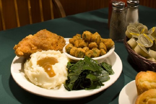 A plate of fried chicken, mashed potatoes with gravy, collard greens, and fried okra, with condiments in the background.