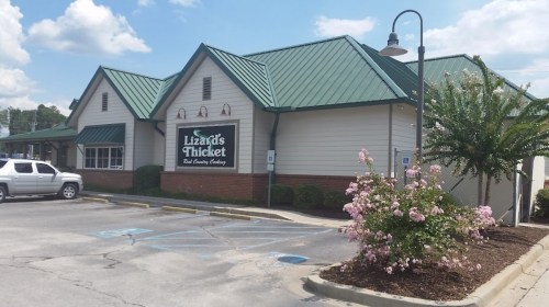 A restaurant building with a green metal roof, sign reading "Lizard's Thicket," surrounded by flowers and parking spaces.