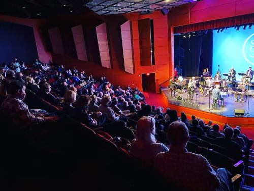 A large audience watches a performance on stage in a well-lit theater with red and blue lighting.