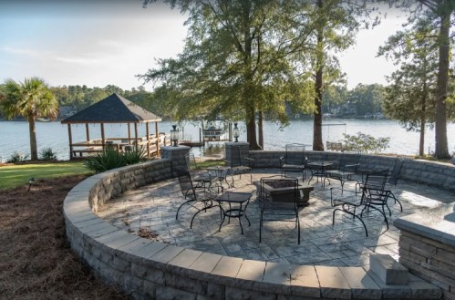 A circular stone patio with chairs surrounds a fire pit, overlooking a lake and gazebo in a serene outdoor setting.