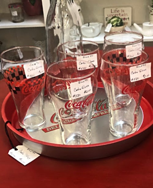 A tray with six Coca-Cola glasses and a bottle, each with price tags, displayed in a shop setting.