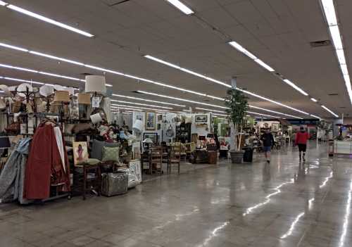 A spacious thrift store aisle filled with various furniture, decor items, and shoppers browsing.