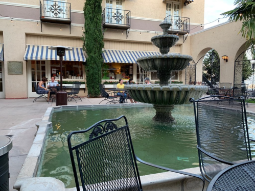 A courtyard with a fountain, surrounded by tables and chairs, and a restaurant in the background.