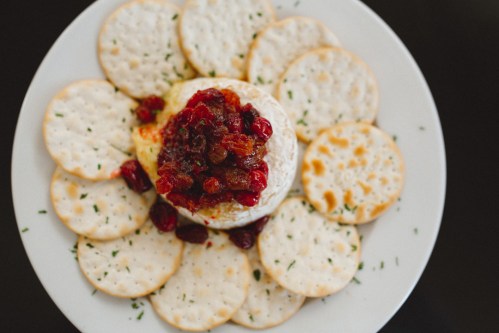 A round cheese wheel topped with cranberry sauce, surrounded by circular crackers on a white plate.