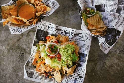A colorful plate of nachos topped with lettuce, tomatoes, and cheese, served with tortilla chips and salsa on newspaper.