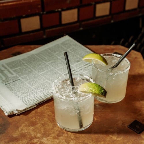 Two cocktails with lime wedges and straws on a wooden table, next to a folded newspaper.