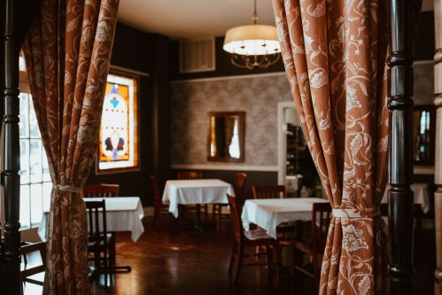 Cozy restaurant interior with tables, elegant curtains, and a stained glass window. Warm, inviting atmosphere.