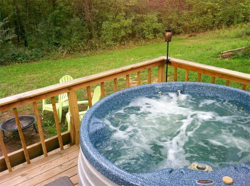 A hot tub on a wooden deck surrounded by greenery, with two Adirondack chairs nearby.