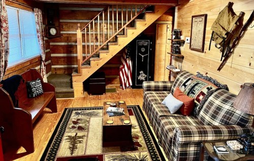 Cozy wooden cabin interior with a plaid sofa, coffee table, and staircase, featuring rustic decor and warm lighting.