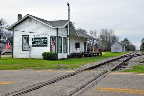 Historic train station with "Jimmy Carter Presidential Center" sign, flags, and railroad tracks in a rural setting.