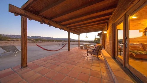 A cozy patio with a hammock, chairs, and a view of mountains at sunset. Warm wooden architecture and tiled flooring.