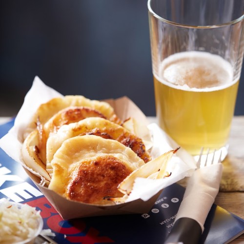 A serving of golden-brown dumplings in a paper tray, accompanied by a glass of beer on a wooden table.