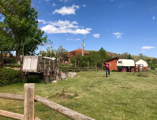 A scenic outdoor view featuring vintage wagons, a red barn, and a person standing in a grassy area under a blue sky.
