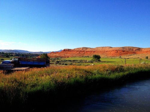 A blue truck parked near a river, with green fields and red rock formations under a clear blue sky.