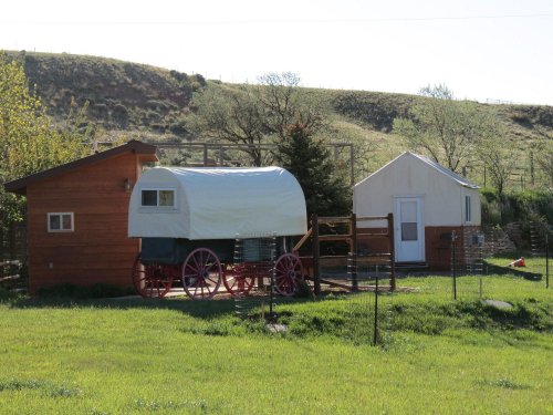 A rustic scene featuring a covered wagon and a small cabin in a grassy area, surrounded by trees and hills.