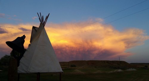 A teepee silhouette against a vibrant sunset sky with colorful clouds and distant hills.