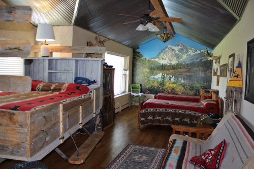 Cozy rustic bedroom with wooden furniture, colorful bedding, and a scenic mural of mountains and a lake.
