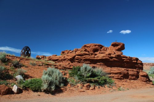 A rocky landscape with red rock formations, green shrubs, and a clear blue sky with a few clouds.