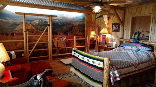 Cozy rustic bedroom with wooden decor, a bed with colorful blankets, and a scenic mountain mural. Warm lighting throughout.