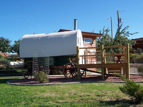 A vintage wagon with a white cover, set on a grassy area next to a wooden structure and surrounded by trees.