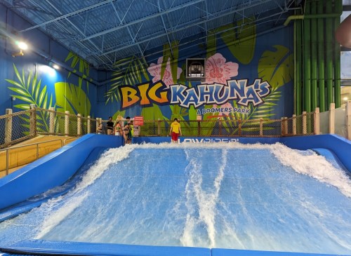 Two people stand on a surf simulator at Big Kahuna's water park, with vibrant tropical-themed walls in the background.