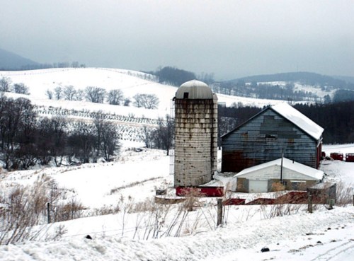 A snowy landscape featuring a barn and silo, with rolling hills and bare trees in the background.