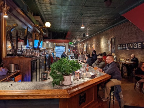 A cozy bar scene with patrons seated at the counter and tables, featuring warm lighting and rustic decor.