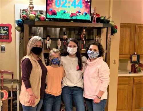 Four people wearing masks stand together in a festive room with holiday decorations and a timer on the TV.