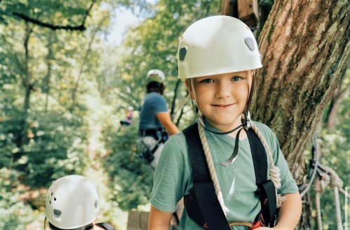 A smiling child in a helmet stands on a tree platform, ready for an outdoor adventure, with others in the background.