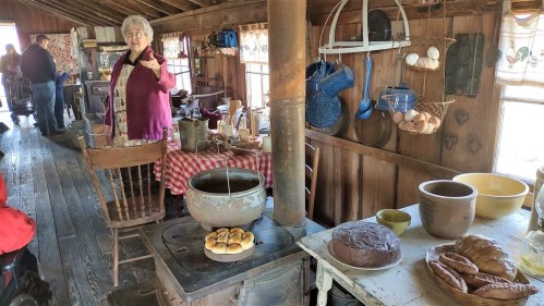 A rustic kitchen with a woman gesturing, featuring a pot on the stove and various baked goods on the table.
