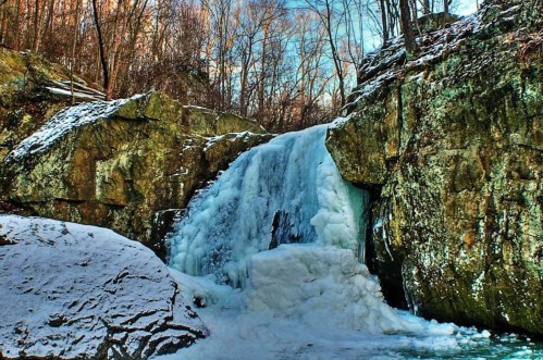 A frozen waterfall cascading over rocky terrain, surrounded by snow-covered ground and bare trees.