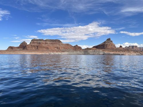 A serene view of red rock formations reflected in calm water under a blue sky with scattered clouds.