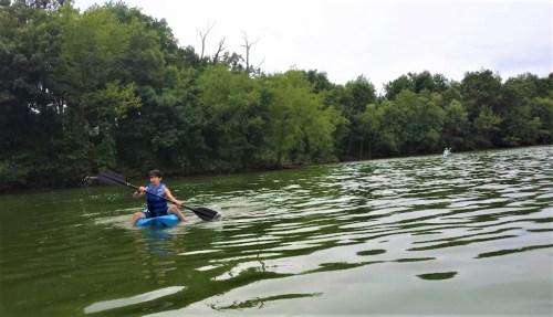 A person paddles a kayak on a calm green lake surrounded by trees under a cloudy sky.
