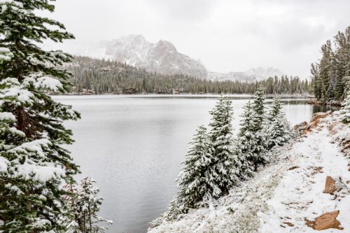 A snowy landscape by a lake, surrounded by evergreen trees and mountains under a cloudy sky.