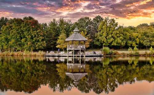 A serene lakeside gazebo at sunset, surrounded by colorful trees and reflecting in the calm water. Birds fly overhead.