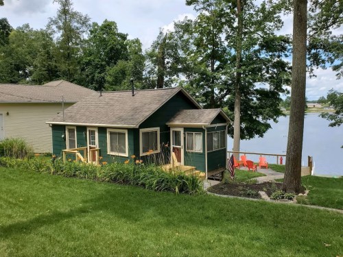 A green cottage by a lake, surrounded by trees and a well-kept lawn, with a patio and orange chairs by the water.